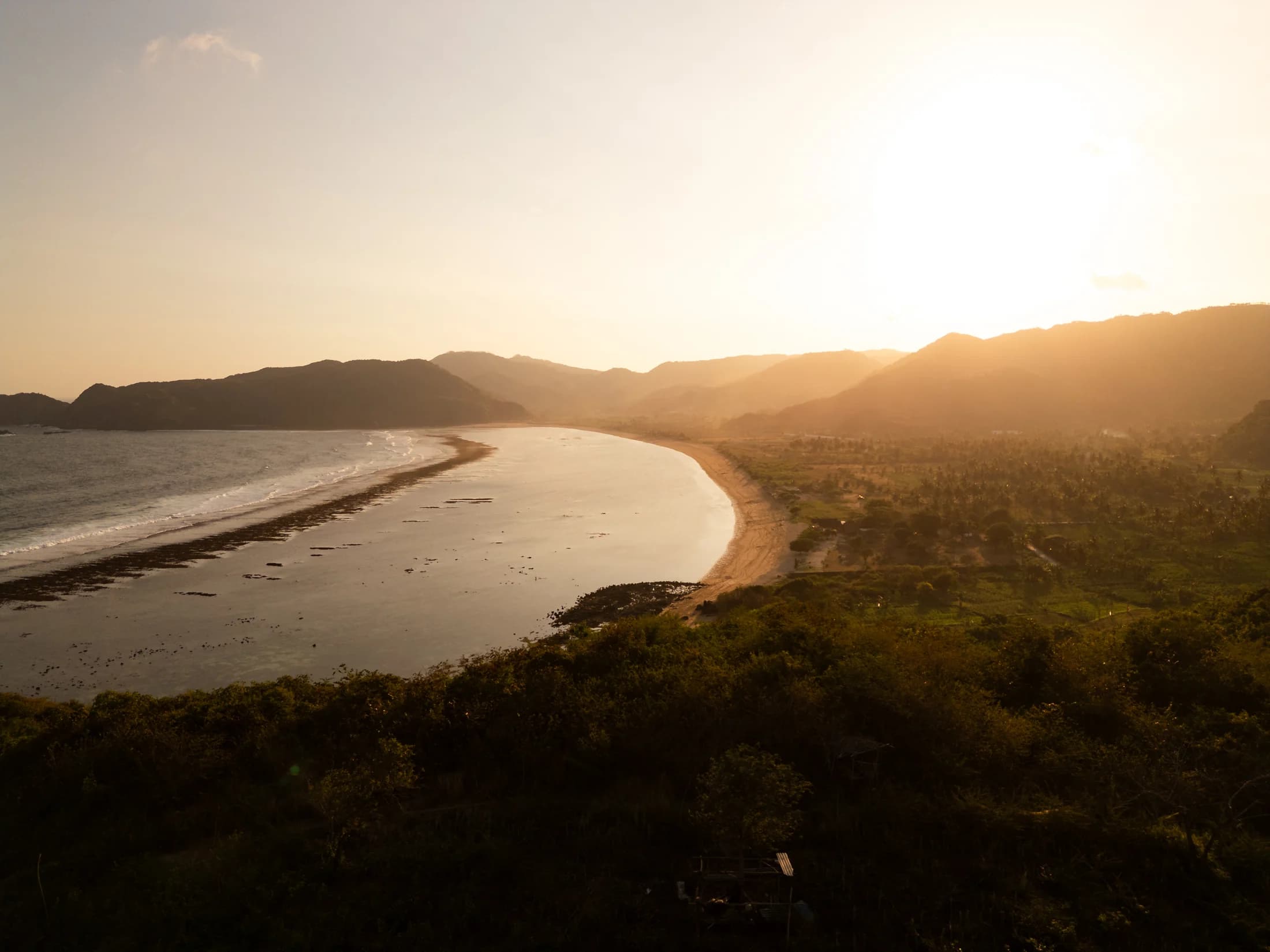 Luchtbeeld van Tampah Bay bij zonsondergang met de gebogen kustlijn en heuvels rond AURAH Lombok.